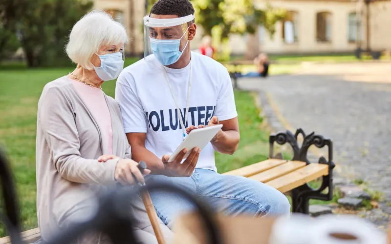 Young male volunteer wearing a protective shield while sitting on a bench with an aged woman